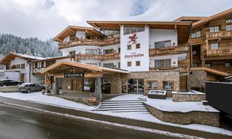 A beautiful hotel with traditional wooden architecture in a wintery setting. The facade is partially covered with snow, surrounded by mountains and trees.