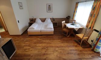 A cozy room with two single beds and a small dining area. The warm wooden floors and the light curtains create an inviting atmosphere.