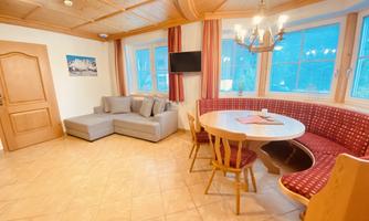 A cozy living space with a couch, a round dining table, and a large window. The room is well-lit and has wooden elements.