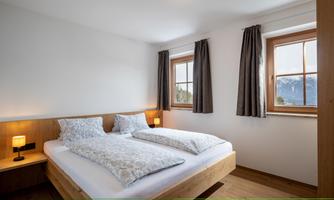 A modern bedroom with a double bed and two windows. The walls are light, and there are wooden details as well as cozy lighting.