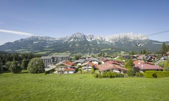 A picturesque landscape with green meadows and small houses. In the background, impressive mountains rise under a clear blue sky.