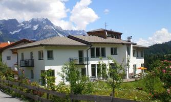A modern house with a balcony, surrounded by green landscape and flowers. In the background, the mountains are visible under a blue sky with some clouds.