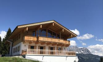 A modern chalet in the mountains with wooden cladding and large windows. In the background, majestic mountains and a clear sky can be seen.