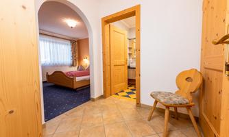 A bright hallway with tiled flooring and wooden doors. In the background, there is a bedroom with a bed and windows visible.