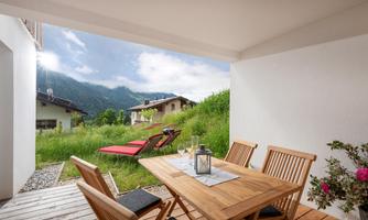 A cozy dining area on a terrace with wooden chairs and a table. In the background, green meadows and mountains can be seen.
