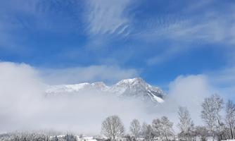 A winter mountain with snow-covered peaks under a blue sky. In the foreground, there are trees and some fog visible.