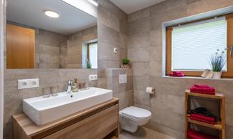 A modern bathroom with a large wooden sink unit. There is a mirror, a toilet, and windows with blinds.