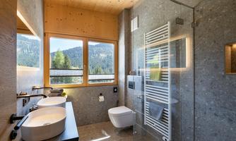 A modern bathroom with large windows and a view of the mountains. It features two sinks and a tiled shower.