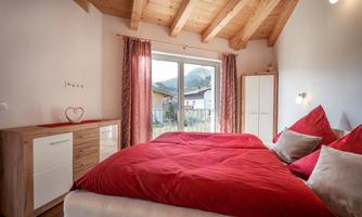 A cozy bedroom with a large bed and red pillows. The large windows offer a view of the landscape.