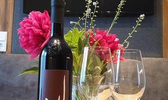 A bottle of wine is on a wooden table, surrounded by two wine glasses and colorful flowers. In the background, a television is visible.