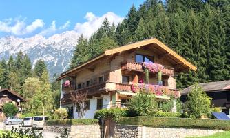 A charming wooden house with balconies, surrounded by lush greenery and a view of the mountains in the background. The facade is adorned with colorful flowers, creating an idyllic atmosphere.