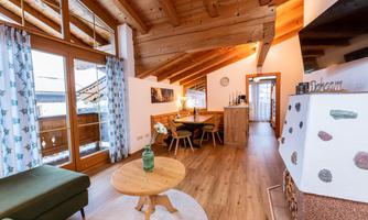 A modern, cozy apartment with a wooden beam ceiling and large windows. The living area is bright and inviting, featuring a dining table and a seating area.