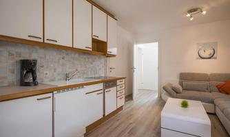 A modern kitchen with white cabinets and a refrigerator. In the living area, there is a gray sofa and a coffee table.