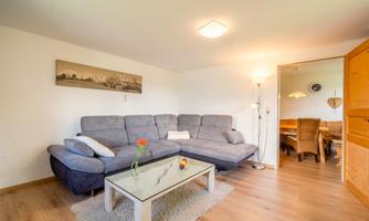 A modern living room with a gray couch and a white table. In the background, a dining area is visible.