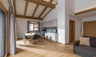 A modern, bright living room with a wooden ceiling and large windows. There is a dining area with chairs and a cozy spot to relax.