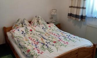 A cozy bedroom with a double bed and colorful bedcovers. The walls are bright and a window with curtains provides daylight.