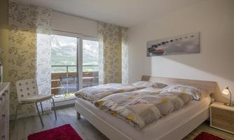 A cozy bedroom with two beds and a large window.  
There are light walls, a chair, and a red carpet.