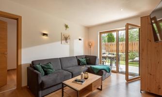 A modern living room with a gray sofa and a wooden table. Large windows offer a view of the garden.