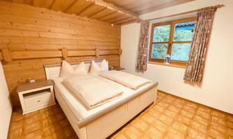 A cozy bedroom with wooden walls and a large window. The bed is furnished with two pillows and a blanket.
