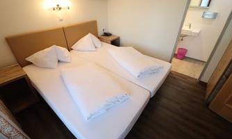 A bright double room with two single beds and fresh bed linen. In the background, a modern bathroom and a television can be seen.