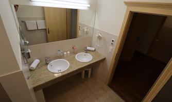 A modern bathroom with two sinks and a large mirror. It is bright and clean, with a wooden door and various hygiene items.
