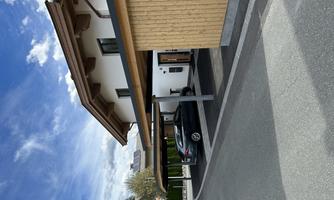 A modern house with wooden cladding and a covered parking space. The sky is blue with some clouds.