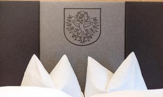 A cozy bedroom with large white pillows and a gray headboard. The headboard features a coat of arms with an eagle.