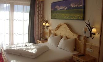 A cozy bedroom with a large bed and light wooden furniture. Sunlight shines through the windows, and a picture of mountains hangs on the wall.