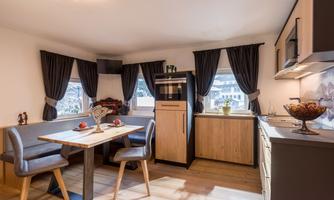 A modern kitchen with a dining area and wooden flooring. The windows are decorated with dark curtains and there are fresh fruits on the table.