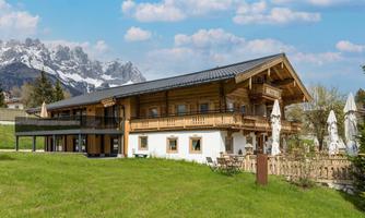 A beautiful wooden house with a modern design, surrounded by green meadows. In the background, majestic mountains and a blue sky can be seen.