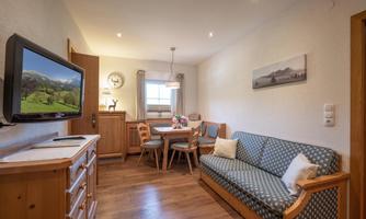 A cozy living space with a sofa, a dining area, and a television. The decor is friendly and modern, featuring wood and neutral tones.