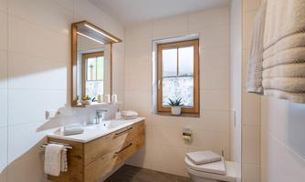 A modern bathroom with wood carvings and a large mirror. There is a bright space with a window view and white tiles.
