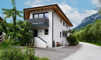 A modern house with a wooden porch and garden, surrounded by trees and mountains. The access road leads to the property.