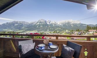 A cozy balcony with a table and chairs, offering a magnificent view of the mountains. The landscape is green and the mountains are snow-covered.