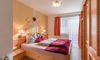 A cozy bedroom with a double bed and colorful cushions. The walls are light and decorated with floral patterns.