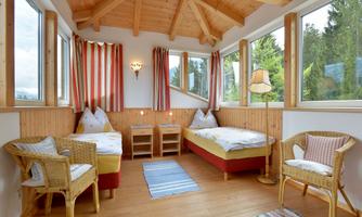 A cozy room with two single beds and a rustic wooden ceiling. Large windows provide plenty of daylight and a view of the greenery.