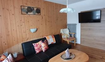 A cozy living room with a black sofa and colorful cushions. Wooden panels are on the wall, and there is a TV as well as a wooden coffee table.