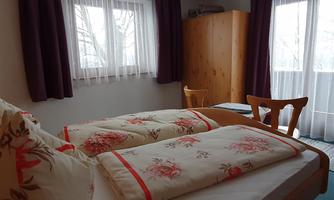 A cozy bedroom with two beds and floral bedding. Large windows let in plenty of light, and a wooden chest of drawers stands against the wall.