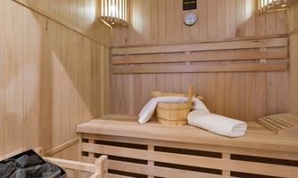 A cozy sauna made of light wood with towels and a bucket. A clock and a heating device are hanging on the wall.
