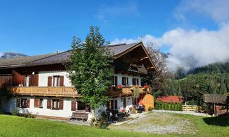 A traditional alpine house with a balcony and colorful flowers. The surroundings are green with mountains and clouds in the background.