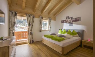 A cozy bedroom with a large double bed and green cushions. The windows offer a view of the mountains and let in plenty of light.