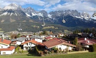 A picturesque landscape with idyllic houses and majestic mountains in the background. Cable cars glide over the scene and the sky is lightly clouded.