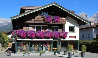 A charming house with a wooden balcony and blooming balcony boxes. In the foreground, shop windows with fashionable clothing can be seen.