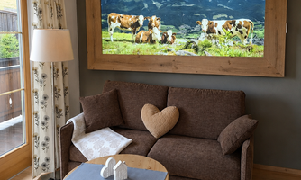 A cozy living room with a brown sofa and a coffee table. On the wall hangs a large picture of cows in a meadow in front of a mountain landscape.