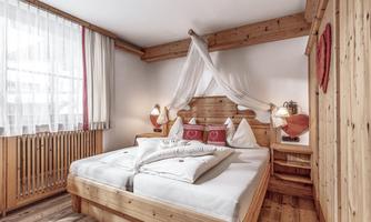 A cozy bedroom with a large wooden bed and white bedding. The room has bright windows and rustic wooden elements.