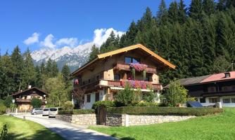 A charming alpine house with a wooden roof and colorful flowers on the balcony. In the background, tall trees and mountains are visible.