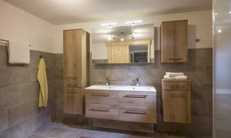 A modern bathroom with wooden shelves and a large mirror. The room is bright and welcoming, featuring gray tiles and a white sink.