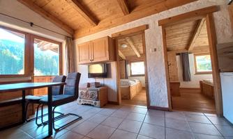 A cozy room with a wooden ceiling and tiled floor. There is a table, chairs, and large windows with a view of the mountains.