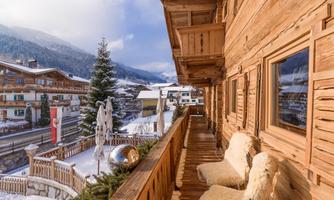 A cozy balcony with wooden furniture and a view of a snowy landscape. In the background, traditional chalets and a beautiful fir tree can be seen.