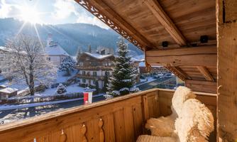 A cozy wooden porch overlooking a snowy landscape. The sun is shining over the snow-covered mountains and houses.